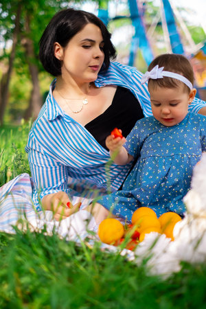 Beautiful mother and her cute daughter eat strawberries and tangerines in the park and enjoy a picnic day, family weekend, park vacationの写真素材