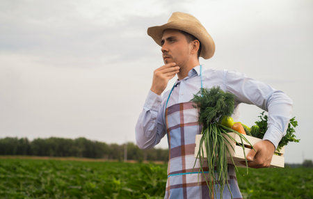 A male organic farmer stands in a vegetable field, holding a wooden box with beautiful freshly picked vegetables, organic vegetables and healthy lifestyle concept.の写真素材
