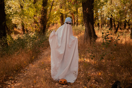 A child in sheets wearing black glasses, looking like a ghost costume in the autumn forest, is...