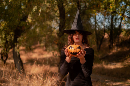 Beautiful woman in witch costume with Jack-o'-lantern in forest, Halloween party, smiling evil witch holding pumpkin and laughing, autumn forest, forest partyの写真素材