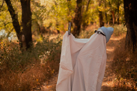 A child in sheets and black glasses, holding a phone in his hands, looking like a ghost costume in...