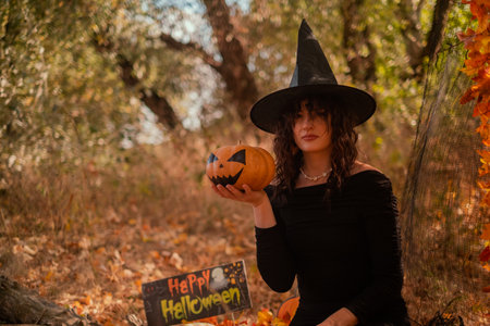 Beautiful woman in witch costume with Jack-o'-lantern in forest, Halloween party, smiling evil witch holding pumpkin and laughing, autumn forest, forest partyの写真素材