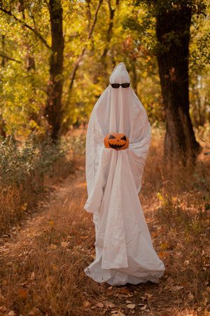 A child in sheets and black glasses, holding a pumpkin in his hands, looking like a ghost costume in the autumn forest, frightening and frightening. Nice little funny ghost. Halloween Partyの写真素材