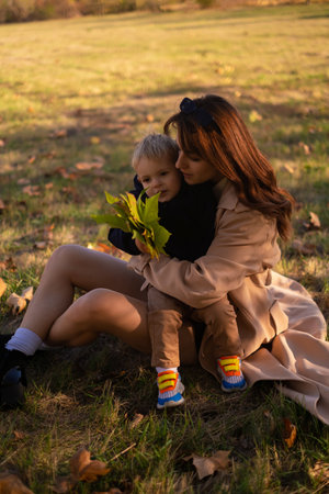 A mother and son embrace and play in the spring park. The boy holds green tree leaves while the mother tickles him, creating precious family moments on Mother's Day, enjoying a picnic in nature.の写真素材
