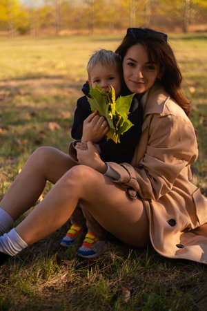 A mother and son embrace and play in the spring park. The boy holds green tree leaves while the mother tickles him, creating precious family moments on Mother's Day, enjoying a picnic in nature.の写真素材