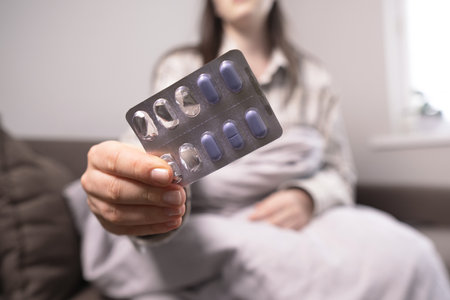 Woman with flu lying under a blanket on the couch, holding a blister pack of antiviral, pain relief, and fever-reducing pills.の写真素材