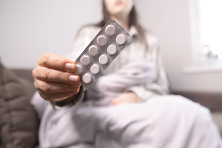 Woman with flu lying under a blanket on the couch, holding a blister pack of antiviral, pain relief, and fever-reducing pills.の写真素材