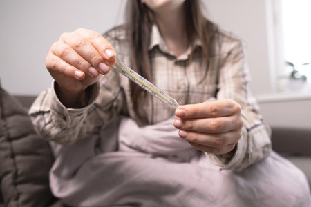 Woman with fever checks her temperature with a thermometer, essential first-aid supplies and home medicine kits nearby.の写真素材