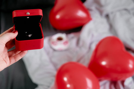 A young woman holds a red ring box with a ring inside, with heart-shaped balloons on the couch in...