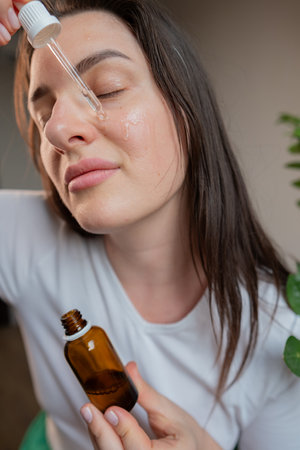 Woman applies facial serum and uses a metal roller for lymphatic drainage and wrinkle smoothing in a skincare routine.の写真素材