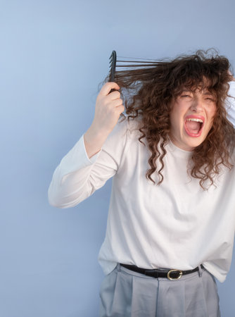 Frustrated curly haired woman screams in pain while trying to comb her tangled hair at home.の写真素材