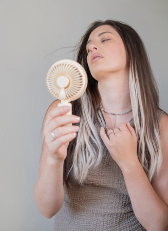 Tired young woman enjoying cool air with a lightweight portable fan outdoors on a warm sunny summer day.の写真素材