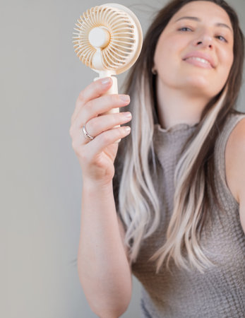Smiling young woman staying cool with a handheld mini fan outdoors on a warm sunny summer day.