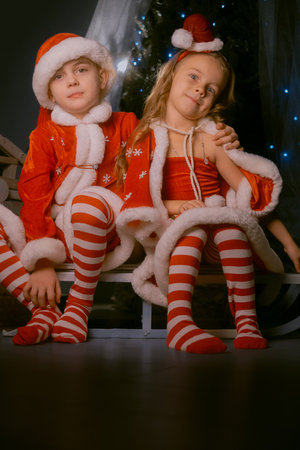 Adorable siblings in festive Santa hats sitting on a sled surrounded by holiday lights and decorations indoors.の写真素材