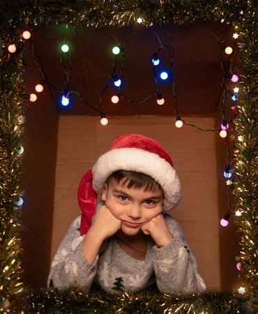 Happy boy sitting in a decorated gift box with lights and tinsel, hands on face, Christmas and New Year moodの写真素材