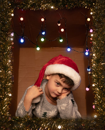 Smiling boy in a festive gift box with sparkling lights and tinsel, hands supporting his face,...