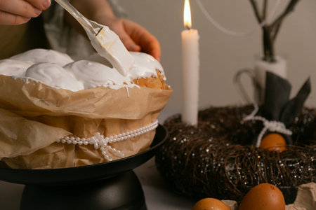 In cozy kitchen, she spreads icing on Easter cake, with eggs, candles, twig wreath on table, and...