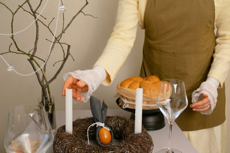 Woman carefully arranging Easter cakes, eggs, and candles on the table, preparing festive treats for...