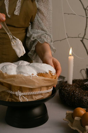 Easter preparation, she spreads white icing on cake, with eggs, candles, twig wreath on table, and...