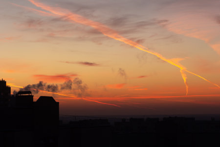 An urban silhouette with a colorful orange and red sky covered with contrails and clouds during sunrise and sunset.の写真素材