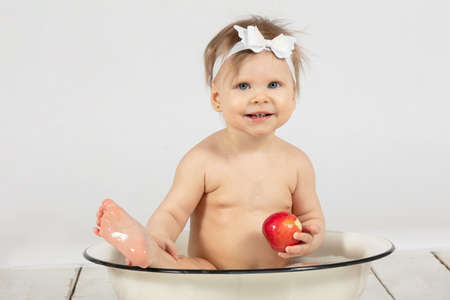 Beautiful baby bathes in a basin and eats a red apple. Little girl in one year old.の写真素材