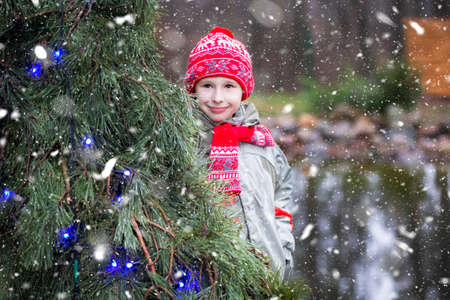 A child on Christmas street. A boy in a red knitted hat stands near a decorated Christmas tree. A...