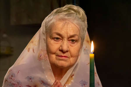 An elderly woman in a headscarf looks at a candle. An elderly woman is looking at a candle. The old woman is praying.の写真素材