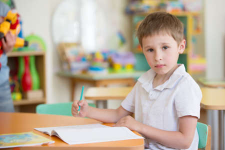 A preschool boy at a table in class with a notebook and penの写真素材