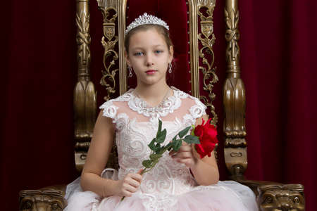 Teenager girl smart in red luxury royal armchair. Girl princess.Queen in a red chair.Luxurious young lady with a scarlet rose in a white dress in an old red armchairの写真素材