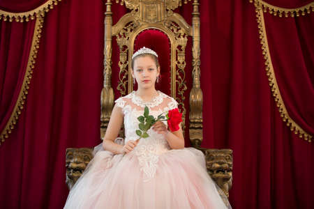 Teenager girl smart in red luxury royal armchair. Girl princess.Queen in a red chair.Luxurious young lady with a scarlet rose in a white dress in an old red armchairの写真素材