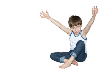 Cheerful preschooler sitting on the floor hands up. Smiling boy in jeans on white background