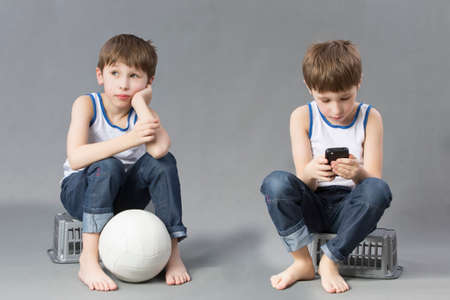 Two brothers twin with different characters. A boy with a soccer ball and his friend with a mobile phone.の写真素材