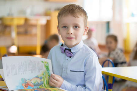 Belarus, the city of Gomel, April 26, 2019. Open day in kindergarten. The boy is a preschooler. Portrait of a six year old child with a book. Junior student at a desk.のeditorial素材