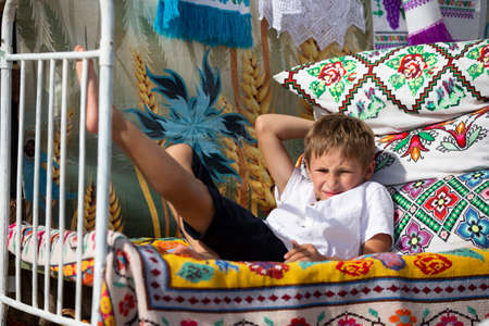 Child Russian or Ukrainian on an old ethnic bed with pillows and bedspreads embroidered with a Slavic pattern. Belarusian boy.の写真素材