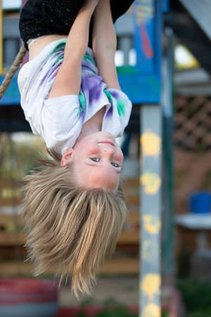 Beautiful little girl hangs upside down on the playground. Little girl smiles into the camera.の写真素材