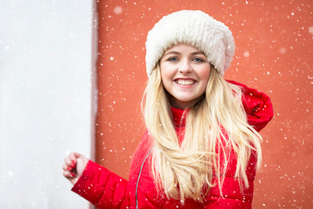 Portrait of a beautiful blonde girl in a warm hat and red jacket and falling snow.の写真素材