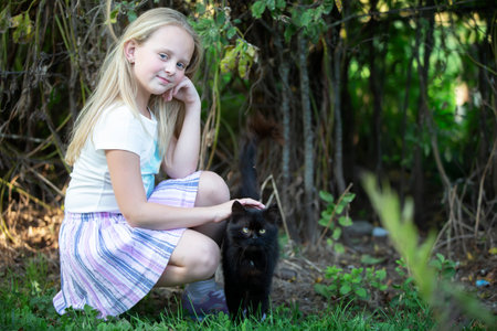 Little blonde girl plays with a black cat on a background of green bushes.の写真素材