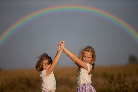 Two little girls stand in a summer field with their hands up under a rainbow. Sisters.の写真素材