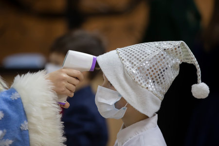 A child wearing a medical mask and a Santa Claus hat is taking the temperature during the coronavirus panademia. Compliance with safety measures for children with coronavirus.の写真素材