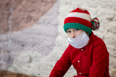 A child in a medical mask wearing a Santa Claus hat due to the coronavirus pandemic. Sad little boy wearing a Christmas hat during the coronavirus epidemic.の写真素材