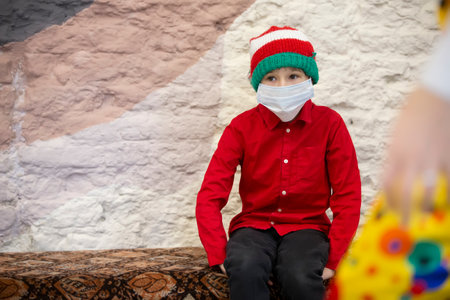 A child in a medical mask wearing a Santa Claus hat due to the coronavirus pandemic. Sad little boy wearing a Christmas hat during the coronavirus epidemic.の写真素材