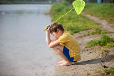 A little boy with a butterfly net sits near the lake and looks at the water.