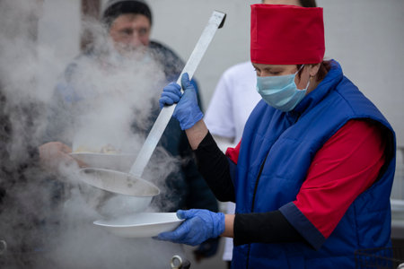 November 13, 2020. Belarus, Gomil region. PA Cooking street food during the coronavirus panademy. A chef in a medical mask with a large scoop pours soup.のeditorial素材