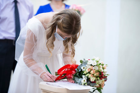 A bride wearing a medical mask with a bouquet signs a marriage certificate during the coronavirus panademy.の写真素材