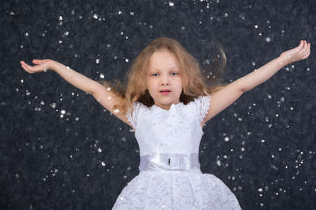 Beautiful little girl in a white dress with raised arms under flying snowflakes.の写真素材