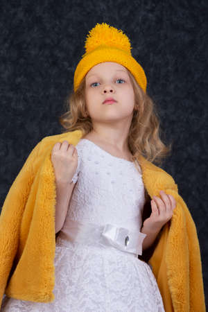 A funny beautiful five-year-old girl in a yellow knitted hat and elegant white dress under white snow.の写真素材