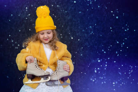 Happy little girl in a yellow fur coat and a knitted hat with skates on a blue background sprinkled with snow.の写真素材