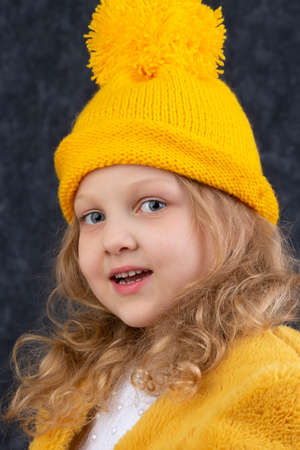 A beautiful little blonde in a knitted yellow hat looks at the camera and smiles. Happy beautiful child about five years old.の写真素材