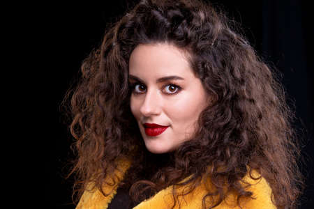 A beautiful brunette girl with curly hair and bright makeup on a dark background looks at the...