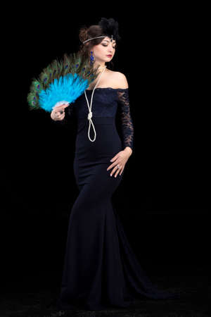 A beautiful girl dressed in the style of the 20-30s of the last century with a fan posing on a dark...